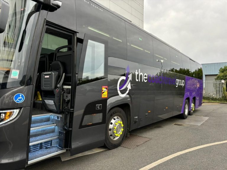 Coach bus with open door featuring "the coach travel group" branding, parked in an industrial area, showcasing accessibility features and modern design.