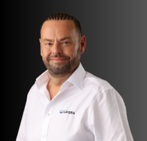 Man with a beard smiling, wearing a white shirt featuring the Layer 4 logo, representing the senior team of Layer 4, a managed IT services provider.